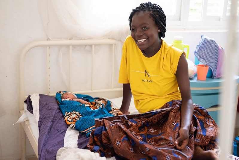 A mother smiling at a camera with her baby laying down on the bed beside her